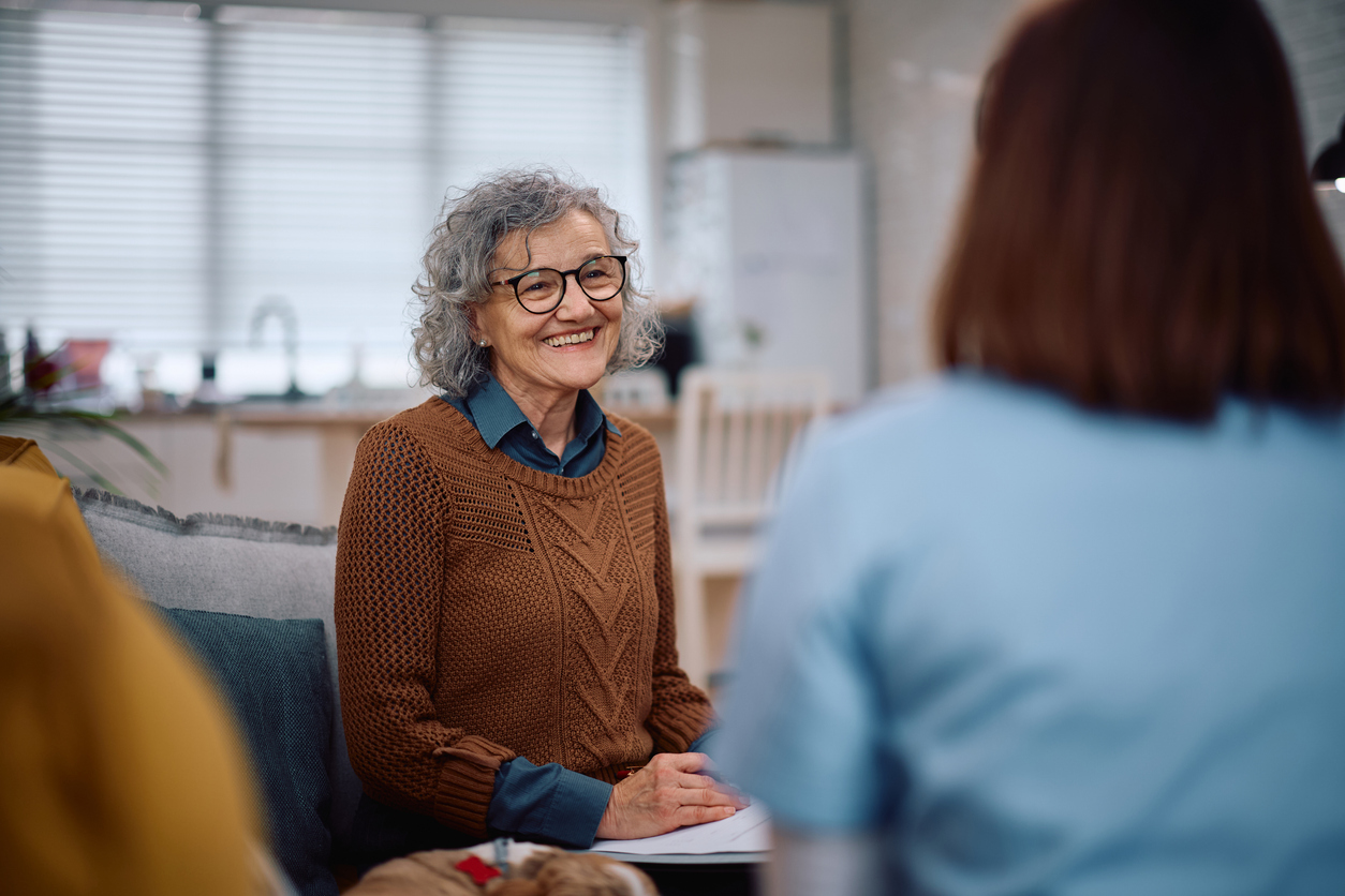 Senior adult woman smiling at caretaker while sitting on couch.