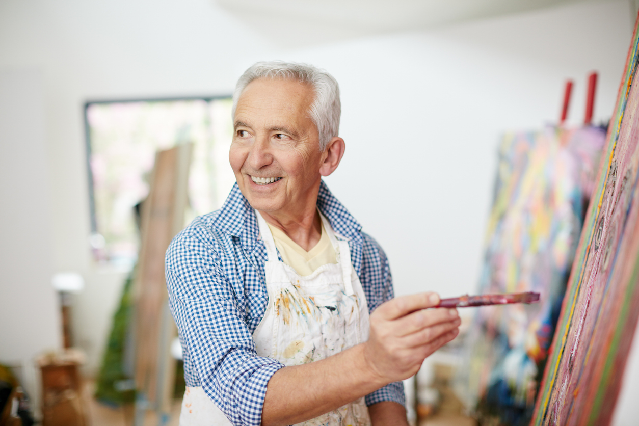 Senior adult man smiles as he paints on canvas.